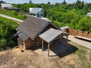 Aerial shot of a traditional wooden hut with a small porch and a wooden fence, surrounded by green hills and other rural buildings in Murom.