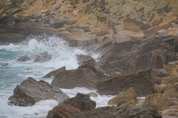 Rocky coast at Praia da Ursa, Sintra, Portugal. Waves, stratified cliffs, erosion, foam, clouds. Geological structure, coastal landscape, natural texture.