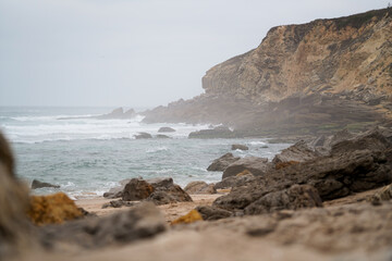 Rocky coast at Praia Pequena do Rodízio, Sintra, Portugal. Waves, stratified cliffs, sand, and clouds. Natural erosion, geological structure, coastal landscape.