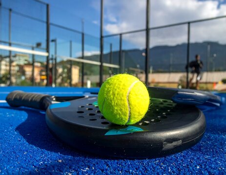 A bright yellow tennis ball rests atop a paddle on a vibrant blue court, with a view of a sports court in the background.