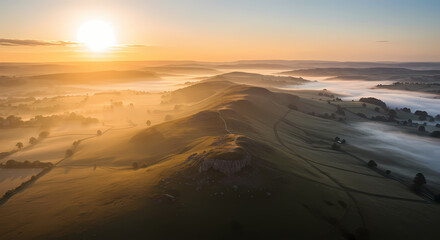 A serene golden sunrise illuminates a misty valley and a long, winding ridge, capturing the tranquil beauty of the rural landscape from an aerial perspective