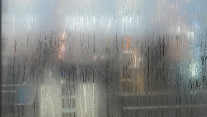 Close-up of wet glass with water droplets, distorting the view of warm glowing lights in the background.