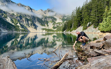 Man kneeling down and touching pristine alpine lake water causing ripples. 