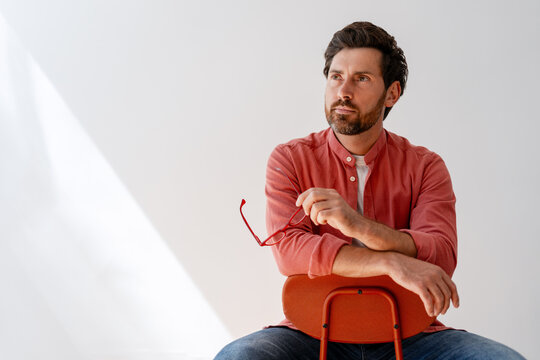 Pensive businessman holding eyeglasses and looking away while sitting on chair - Powered by Adobe