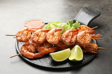 Skewers with tasty grilled shrimps, lime slices, lettuce and sauce on grey table. Delicious seafood dish
