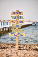 A pole with wooden signs with the text " Please Leave Nothing But Your " against a blurred wooden pier  extending into the sea. Red Sea, Egypt.  Shallow depth of field.