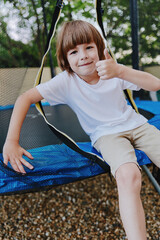 Happy boy giving a thumb up while sitting on a trampoline, wearing a white shirt, enjoying a sunny day outdoors with a playful atmosphere. © SHOTPRIME STUDIO