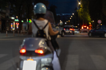 night scene of a young woman on a scooter, wearing a helmet and backpack, stopped at a crosswalk...