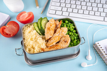 Packed homemade lunch for office, balanced healthy brunch with grilled chicken fillet, couscous garnish, vegetables, in lunch box, with keyboard, office accessories, healthy eating at work meal prep