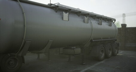 A large tanker truck stands parked in a dimly lit area, surrounded by fog. The metallic surface glistens as the mist envelops the environment, creating an industrial atmosphere.