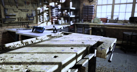 A well equipped workshop is illuminated by natural light streaming through large windows. The focal point is a rugged workbench scattered with sawdust and surrounded by various hand tools.