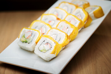 A row of sushi rolls with a yellow outer layer, likely tamago, filled with crab and avocado. The rolls are elegantly presented on a long, modern white plate.
