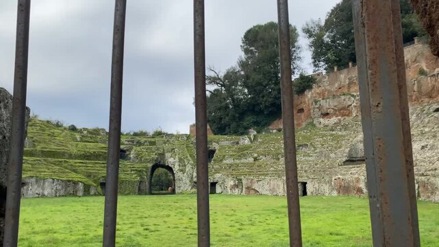 Dolly out shot of closed metal gate and old ruined arena stage for battles overgrown with greenery, abandoned amphitheatre being a part of medieval history, cultural monument seen through the gateway