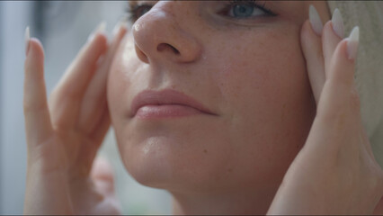 A young woman gently massages her face while applying a serum in a bright bathroom, focusing on her skincare regimen.