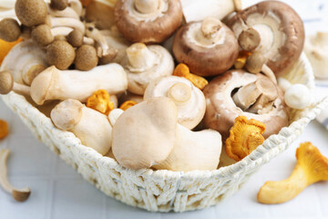 Different raw mushrooms in wicker basket on white tiled table, closeup