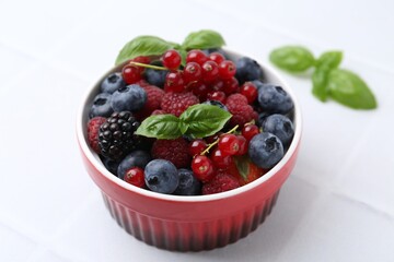 Different ripe berries and basil leaves in bowl on white tiled table, closeup