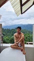 Shirtless man relaxing by an outdoor bathtub with a scenic forest and mountain backdrop on a transparent background