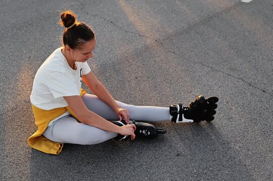 A young woman sitting on the asphalt is adjusting her inline roller skates. The image captures a moment of preparation or a break from the sport and represents an active and healthy lifestyle.