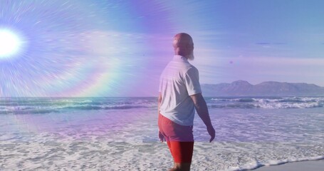 Gazing man wearing white T-shirt red trunks wading beach surf, with sun flare mountains, copy space