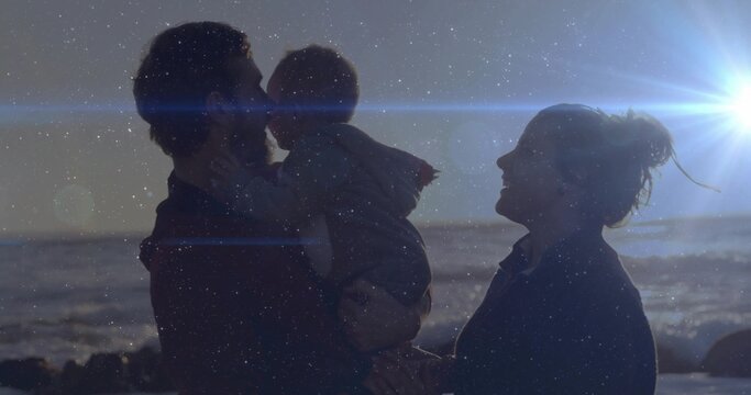 Father wearing jacket holding baby, kissing him with smiling mother at shoreline, ocean lens flare - Powered by Adobe