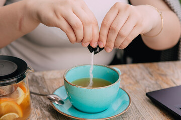 Close-up: woman's hands and the moment of sweetening herbal tea . Glass teapot with fruits on wooden cafe table .