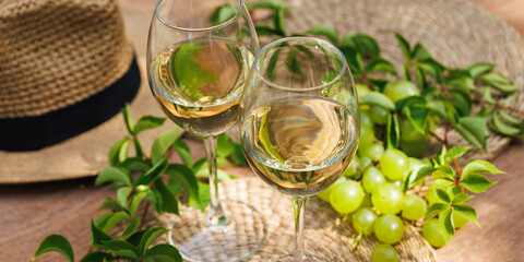 Overhead view of two white wine glasses with green grapes, vines and hat on summer table..