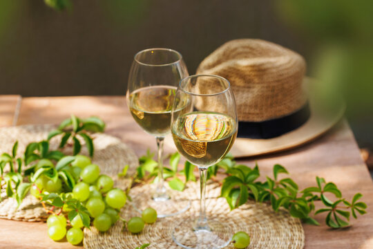 Two glasses of white wine with green grapes and straw hat on wooden patio table in natural light..