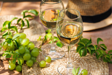 Two wine glasses with white wine near fresh grapes, leafy greens and straw hat on wooden surface..
