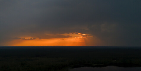 Golden sunlight breaks through dark clouds at sunset over a vast forest landscape, creating a dramatic contrast between light and shadow, showcasing nature's beauty.