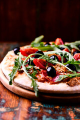 Pizza with salami, tomatoes and mozzarella cheese, black olives and arugula. Rustic wooden background. Soft focus. Copy space.	