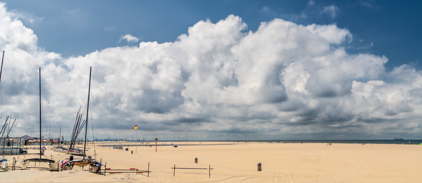 Catamarans and sailboats lie anchored on a vast, sandy beach under a sky filled with large, fluffy clouds. The expansive Kijkduin beach of The Hague stretches out to meet the horizon.