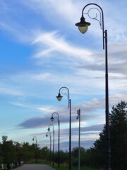 Graceful row of decorative street lamps in a park, rising elegantly against pastel blue sky with delicate white cloud streaks, creating a serene urban landscape.
