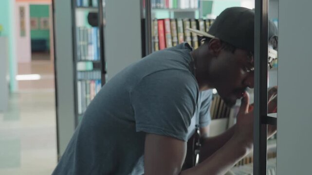 Young man in casual grey shirt and backwards cap squatting near library shelf, browsing and examining books with thoughtful expression, surrounded by colorful book spines in quiet indoor setting