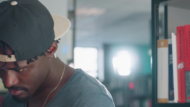 Close up side view of young man in casual clothing and backward cap standing near bookshelf, gazing downward with serious contemplative expression
