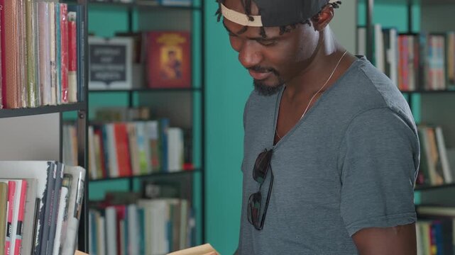 Young man in casual clothing and backward cap standing near bookshelves in library, holding open book and reading attentively, surrounded by colorful spines