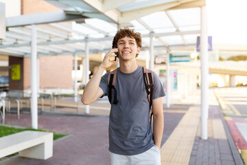 Fototapeta premium Young Caucasian man walking at airport while talking on phone and smiling. Represents casual travel lifestyle, communication, and happy youth on the go