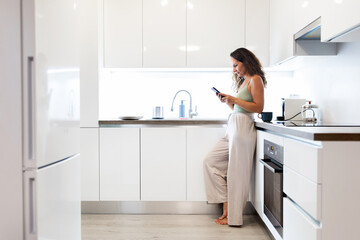 Young Caucasian woman at home kitchen leaning on counter while using smartphone. Modern domestic lifestyle with relaxation, digital connection and casual routine