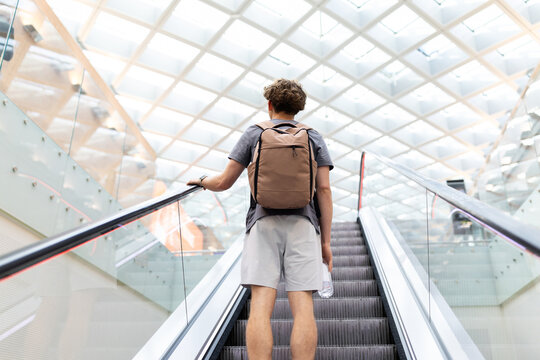 Young Caucasian man with backpack holding water bottle on airport escalator. Represents travel routine, youth journey lifestyle, mobility and passenger waiting - Powered by Adobe