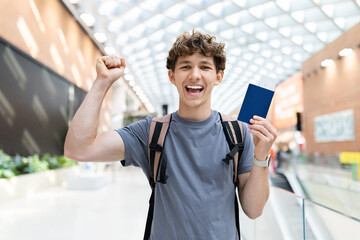 Young Caucasian man with backpack cheering and raising fist while holding passport at airport. Symbol of success, joy, travel excitement and positive lifestyle