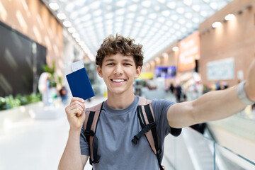 Young Caucasian man taking selfie with passport and boarding pass at airport. Symbol of positive travel lifestyle, departure routine, youth energy and happiness