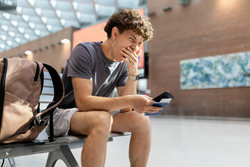 Young Caucasian man sitting at airport bench yawning tired with passport and smartphone. Represents fatigue, waiting stress, flight delay, and youth travel lifestyle