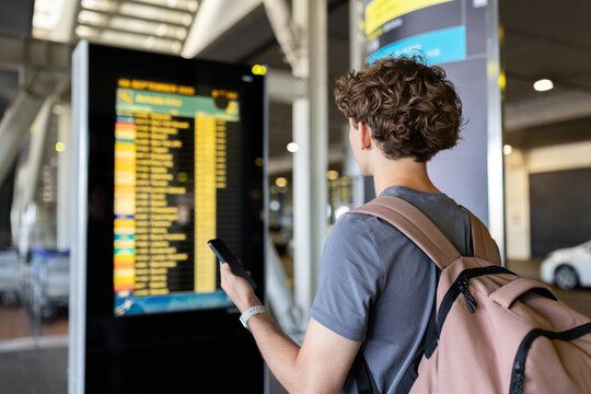 Young Caucasian man with backpack checking flight information board at airport. Travel concept with waiting, flights, passenger journey and digital mobility