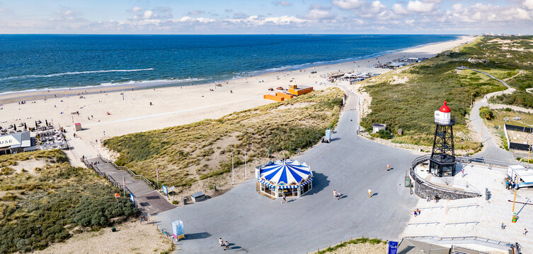 A panoramic view of a sandy beach of Kijkduin with a clear view of the sea. A lighthouse and a carousel are prominent near the shore, surrounded by pathways and greenery.