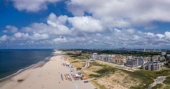 A panoramic view of a beach stretching along the city of The Hague. The sandy coastline meets the deep blue sea. At the horizon on the right, the high-rise buildings of the city center are visible