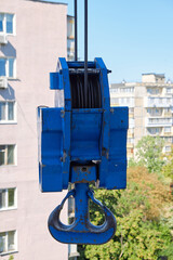Close-up of a blue industrial crane hook with metal cable against a clear sky and residential building background. Heavy machinery detail used in construction and lifting work