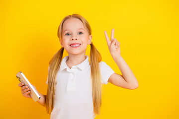 Happy young girl holding a phone and showing a peace sign on a vibrant yellow background, charming...