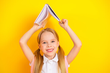 Smiling schoolgirl holding a book on her head against a vibrant yellow background emphasizing...