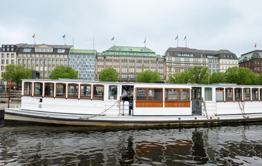 Naklejka premium Vintage passenger ship on the Alster river, with multi-story buildings and trees in the background. Urban waterfront scene.