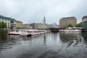 Naklejka premium Hamburg, Germany – Tour boats on the Binnenalster in front of the historic City Hall on a cloudy day. Reflections in the water and urban architecture.