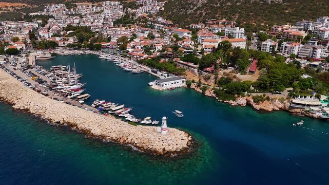 Drone aerial view of Kas marina in Turkey, featuring a stone breakwater with a lighthouse, moored yachts, boats, and the coastal town with hillside houses in the background.